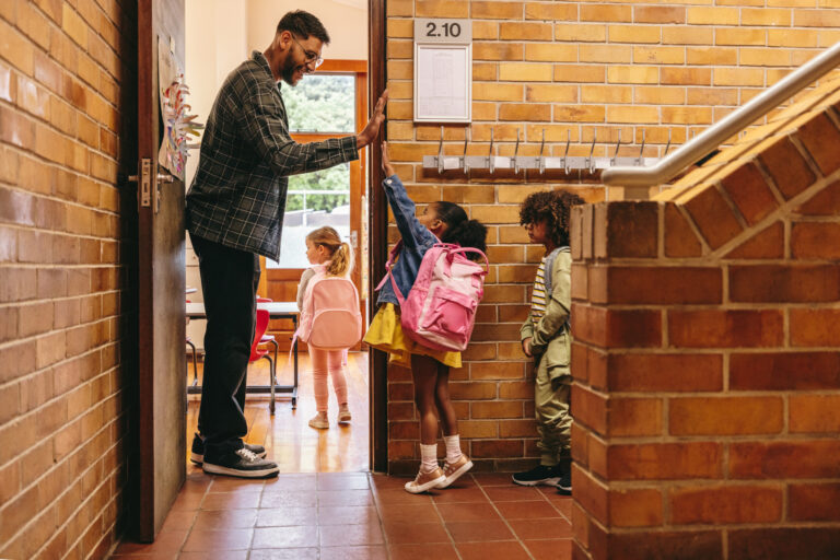 Elementary school kindergarten teacher greeting his students at the door. Male teacher welcoming his class with a high five outside their classroom. Child mentor motivates his pupils in a primary school.